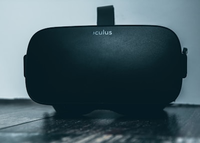 A black virtual reality headset with the brand name 'Oculus' is resting on a dark wooden surface. The headset is positioned upright with the lens end facing towards the viewer, and the background is a plain light-colored wall.