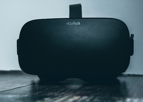 A black virtual reality headset with the brand name 'Oculus' is resting on a dark wooden surface. The headset is positioned upright with the lens end facing towards the viewer, and the background is a plain light-colored wall.