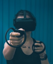 A person wearing a virtual reality headset and holding motion controllers in both hands. The background features vertical blue panels, creating a sleek and modern look. The focus is on the outstretched hand with the controller, suggesting interaction with a virtual environment.