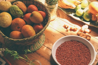 A woven basket filled with various fruits including apples, lemons, and mangoes sits on a wooden table. Nearby, there's a bowl of red seeds or grains and a variety of other fruits like bananas and oranges displayed on a platter. Fresh ginger root and some green herbs can also be seen.