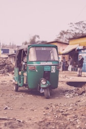 A green three-wheeled auto rickshaw is parked on a dirt road surrounded by debris and sparse vegetation. In the background, a couple of buildings and a person can be seen, creating an urban, slightly rural setting.