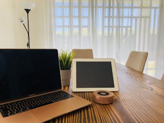 A modern workspace setup on a wooden table featuring a closed laptop, a tablet, a cylindrical speaker, and a small potted plant. The room is well-lit with natural light streaming through sheer window curtains. A tall floor lamp stands in the corner, and cushioned chairs are arranged around the table.
