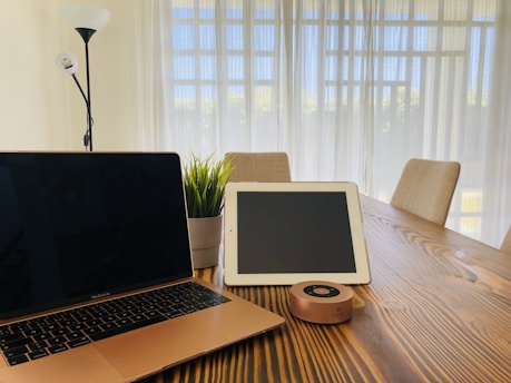 A modern workspace setup on a wooden table featuring a closed laptop, a tablet, a cylindrical speaker, and a small potted plant. The room is well-lit with natural light streaming through sheer window curtains. A tall floor lamp stands in the corner, and cushioned chairs are arranged around the table.