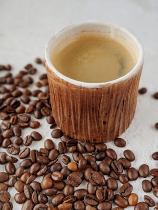 A rustic wooden cup filled with creamy coffee is surrounded by scattered coffee beans on a light surface.