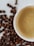A white ceramic cup filled with creamy coffee sits on a surface scattered with roasted coffee beans. The coffee has a smooth surface with a few bubbles, indicating freshness.
