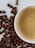 A white ceramic cup filled with creamy coffee sits on a surface scattered with roasted coffee beans. The coffee has a smooth surface with a few bubbles, indicating freshness.