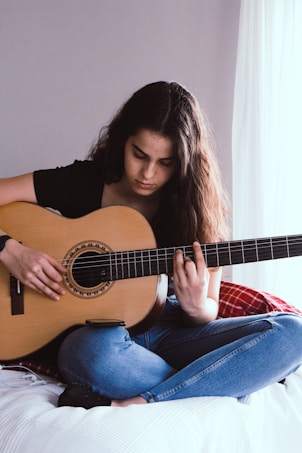 A person sitting cross-legged on a bed playing an acoustic guitar, focused on their music. The room has a soft ambiance with natural light filtering through a sheer curtain.