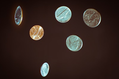 Various coins appear to be suspended or floating against a dark background. The coins are in different positions, showcasing both sides and edges, with some showing clear engravings while others have a more blurred appearance.