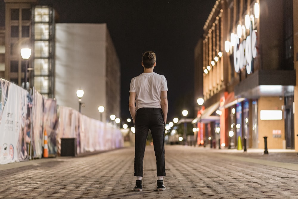 man in white crew neck shirt and black pants standing on sidewalk during night time