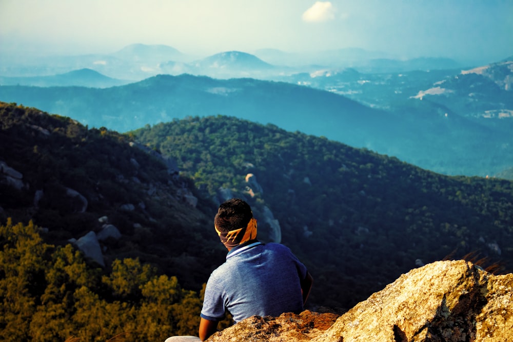 man in blue t-shirt sitting on brown rock looking at the mountains during daytime