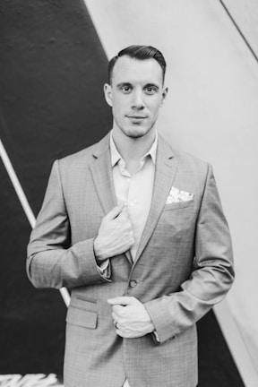 A man is wearing a light-colored suit with a patterned pocket square, standing against a backdrop featuring diagonal lines. He is looking directly at the camera with a gentle expression, with one hand touching the lapel of his jacket and the other positioned near his waist.