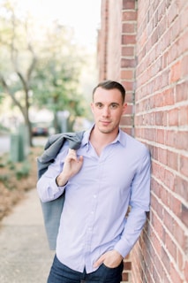 A man wearing a casual jacket and jeans leaning against a rustic brick wall
