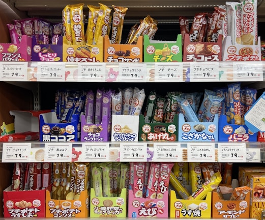 A variety of packaged snacks and treats are neatly displayed on grocery store shelves. The packaging features vibrant colors such as red, yellow, and blue, with labels in Japanese text. Each row on the shelves showcases different types of snacks, including both sweet and savory options.