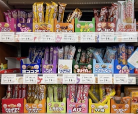 A variety of packaged snacks and treats are neatly displayed on grocery store shelves. The packaging features vibrant colors such as red, yellow, and blue, with labels in Japanese text. Each row on the shelves showcases different types of snacks, including both sweet and savory options.