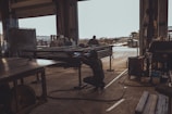 A person is welding metal inside a dark, industrial workshop, with sunlight streaming through an open door. The space is filled with various pieces of equipment and metal structures. Another individual is visible in the background working on a similar task, while the outside area appears to be an industrial yard.