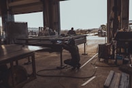 A person is welding metal inside a dark, industrial workshop, with sunlight streaming through an open door. The space is filled with various pieces of equipment and metal structures. Another individual is visible in the background working on a similar task, while the outside area appears to be an industrial yard.