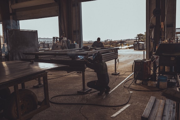 A person is welding metal inside a dark, industrial workshop, with sunlight streaming through an open door. The space is filled with various pieces of equipment and metal structures. Another individual is visible in the background working on a similar task, while the outside area appears to be an industrial yard.