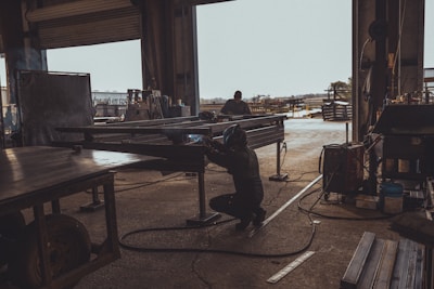 A person is welding metal inside a dark, industrial workshop, with sunlight streaming through an open door. The space is filled with various pieces of equipment and metal structures. Another individual is visible in the background working on a similar task, while the outside area appears to be an industrial yard.