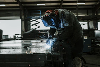 A skilled welder in protective gear carefully joining metal pieces in a bright, modern workshop filled with industrial tools.
