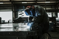 A welder wearing protective gear is working on a metal piece in a dimly lit industrial workshop. Sparks fly from the welding torch, casting light on the surrounding area.