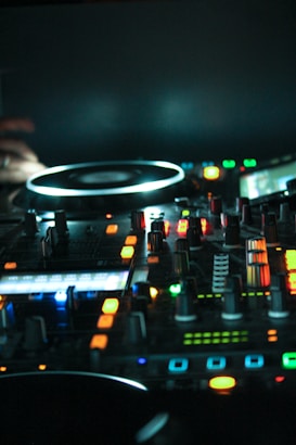 A DJ controller with various buttons, knobs, and lights, predominantly illuminated in blue and orange hues. The scene is dimly lit, creating a dynamic and high-tech ambiance.