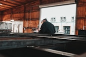 A person is welding metal inside a large industrial workshop. The space is dimly lit with wooden walls and a partially open large garage door. Sparks fly from the welding process, illuminating the immediate area around the worker.
