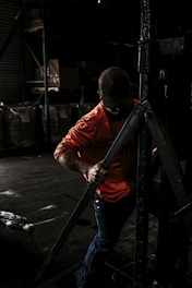 A person wearing a bright orange shirt and a cap is working in a dimly lit industrial or warehouse setting, handling a large metallic structure or equipment. The background is filled with shelves and large bags or boxes.
