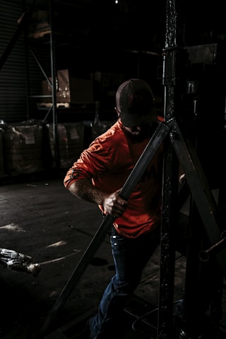 A person wearing a bright orange shirt and a cap is working in a dimly lit industrial or warehouse setting, handling a large metallic structure or equipment. The background is filled with shelves and large bags or boxes.