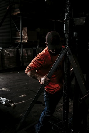 A person wearing a bright orange shirt and a cap is working in a dimly lit industrial or warehouse setting, handling a large metallic structure or equipment. The background is filled with shelves and large bags or boxes.