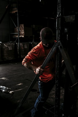 A person wearing a bright orange shirt and a cap is working in a dimly lit industrial or warehouse setting, handling a large metallic structure or equipment. The background is filled with shelves and large bags or boxes.