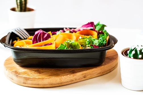 A vibrant salad with colorful vegetables such as orange carrot ribbons, purple cabbage, and green lettuce in a black rectangular container. It sits on a round wooden board. The background includes two potted cacti in white pots, adding a fresh and natural feel to the scene.