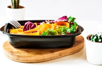 A vibrant salad with colorful vegetables such as orange carrot ribbons, purple cabbage, and green lettuce in a black rectangular container. It sits on a round wooden board. The background includes two potted cacti in white pots, adding a fresh and natural feel to the scene.