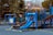 A playground features a variety of equipment, including a prominent blue slide and several platforms connected by steps and railings. The area is surrounded by tall trees and an American flag is visible atop one of the structures. Yellow caution tape is hung around parts of the playground, indicating it may be temporarily closed or under maintenance.