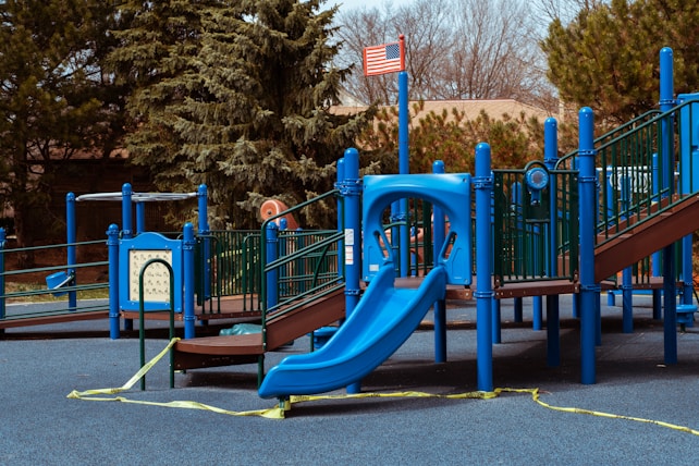 A playground features a variety of equipment, including a prominent blue slide and several platforms connected by steps and railings. The area is surrounded by tall trees and an American flag is visible atop one of the structures. Yellow caution tape is hung around parts of the playground, indicating it may be temporarily closed or under maintenance.