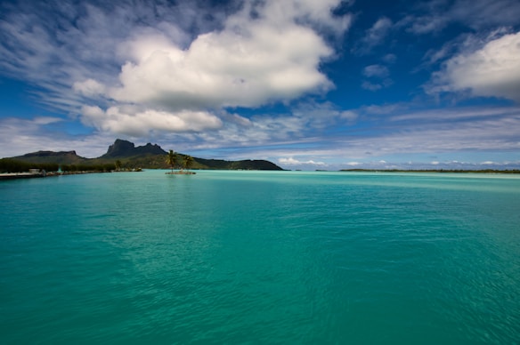 A vivid tropical seascape featuring calm turquoise waters stretching towards a distant island with lush greenery and jagged mountain peaks under a partly cloudy sky.