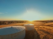 Two large, cylindrical water storage tanks are situated in a field of dry, golden grass. The sun is setting or rising in the background, casting a warm light across the landscape. Trees and other vegetation can be seen in the distance under a clear blue sky.