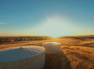 Two large, cylindrical water storage tanks are situated in a field of dry, golden grass. The sun is setting or rising in the background, casting a warm light across the landscape. Trees and other vegetation can be seen in the distance under a clear blue sky.