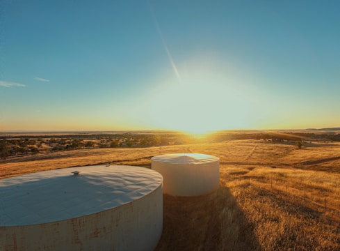 Two large, cylindrical water storage tanks are situated in a field of dry, golden grass. The sun is setting or rising in the background, casting a warm light across the landscape. Trees and other vegetation can be seen in the distance under a clear blue sky.