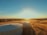 Two large, cylindrical water storage tanks are situated in a field of dry, golden grass. The sun is setting or rising in the background, casting a warm light across the landscape. Trees and other vegetation can be seen in the distance under a clear blue sky.