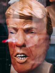 A large mask of a person's face with exaggerated features, including distinct hair and expression. The surface has a textured appearance, suggesting a rubber or plastic material. The mask is displayed behind a reflective glass.
