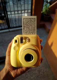 A person holds a yellow Instax Mini 8 camera with a developed photo emerging from the top. The photo features a doodle of various cartoonish faces and shapes. The background includes part of an outdoor setting with a potted plant and a metal gate casting shadows.