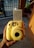 A person holds a yellow Instax Mini 8 camera with a developed photo emerging from the top. The photo features a doodle of various cartoonish faces and shapes. The background includes part of an outdoor setting with a potted plant and a metal gate casting shadows.