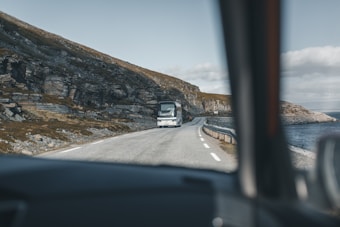 A bus is driving on a narrow coastal road surrounded by rocky cliffs on the left and a body of water on the right. The scene is viewed from inside a car, with the frame partially visible, conveying a sense of movement and anticipation on a road trip.