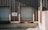 Two closed industrial roller shutter doors are mounted on a weathered, corrugated metal building. A notice sign is attached to the wall between the doors. The area shows signs of rust and aging, particularly on the lower part of the structure. The shadows cast indicate bright sunlight.