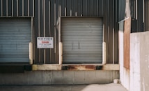 Two closed industrial roller shutter doors are mounted on a weathered, corrugated metal building. A notice sign is attached to the wall between the doors. The area shows signs of rust and aging, particularly on the lower part of the structure. The shadows cast indicate bright sunlight.