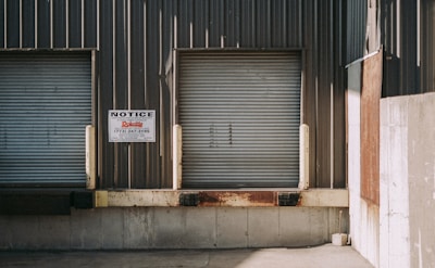 Two closed industrial roller shutter doors are mounted on a weathered, corrugated metal building. A notice sign is attached to the wall between the doors. The area shows signs of rust and aging, particularly on the lower part of the structure. The shadows cast indicate bright sunlight.