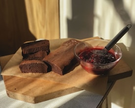 A rustic wooden board holding a loaf of freshly baked chocolate cake, partly sliced, sitting on parchment paper. Beside it is a glass bowl filled with a rich red berry sauce and a whisk resting inside.