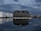 A large industrial building with the name 'Behala' is reflected in a calm body of water. The sky is filled with dark, moody clouds. To the left, there are several large white storage tanks with the logo 'Unitank' visible. On the right, there are industrial cranes and various structures, suggesting a port or dock area. The lighting is dim, suggesting evening or early morning.