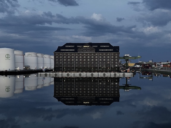 A large industrial building with the name 'Behala' is reflected in a calm body of water. The sky is filled with dark, moody clouds. To the left, there are several large white storage tanks with the logo 'Unitank' visible. On the right, there are industrial cranes and various structures, suggesting a port or dock area. The lighting is dim, suggesting evening or early morning.