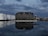 A large industrial building with the name 'Behala' is reflected in a calm body of water. The sky is filled with dark, moody clouds. To the left, there are several large white storage tanks with the logo 'Unitank' visible. On the right, there are industrial cranes and various structures, suggesting a port or dock area. The lighting is dim, suggesting evening or early morning.